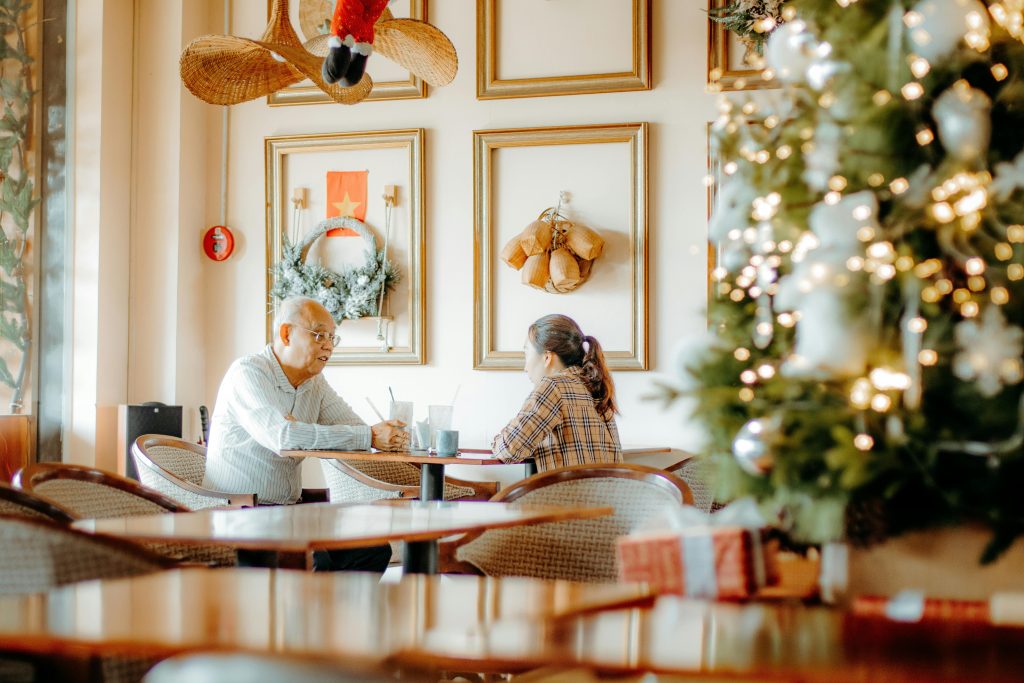 An elderly asian couple sits together at a dining table in a retirement community dining room, with a lit Christmas tree in the foreground.